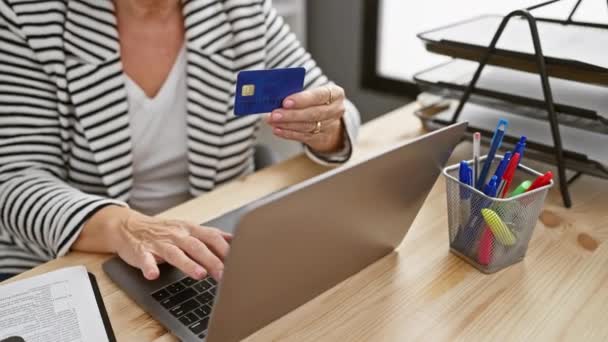 Az érett nő laptoppal vizsgálja meg a hitelkártyát az irodai asztalnál, kifejezve a várakozást és a pénzügyi felelősséget. - Felvétel, videó