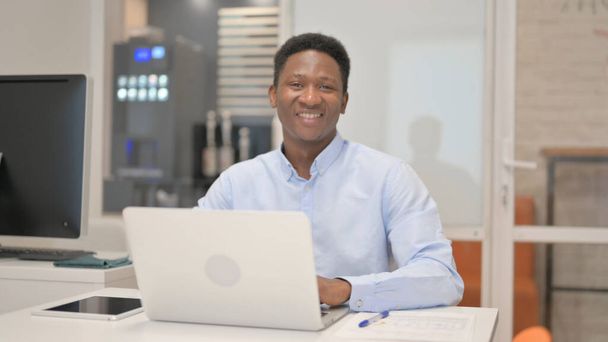 African Businessman uśmiecha się do kamery podczas pracy na laptopie w biurze - Zdjęcie, obraz