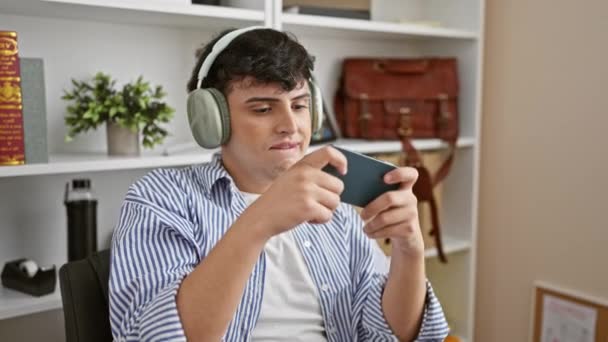 Jeune homme concentré en chemise rayée utilisant un smartphone avec écouteurs dans un bureau moderne. - Séquence, vidéo