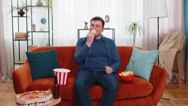 Счастливый кавказский молодой человек в повседневной одежде сидит на удобном диване и ест чипсы, пиццу, попкорн и пиво, смотрит фильмы о спорте, наслаждаясь выходными дома. Улыбающийся парень ест нездоровую пищу на диване в доме. - Кадры, видео