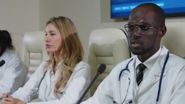 Black doctor in medical coat sitting with committee at press conference table and giving speech in microphone during healthcare event - Footage, Video