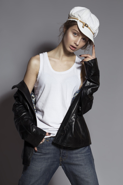 High fashion look. Portrait of a fashionable model with natural  make up and perfect skin, dressed in men's jeans, white shirt, black jacket and stylish white hat.  Studio shot - Zdjęcie, obraz