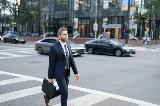 Biznesmen z plecakiem spacerujący po mieście. Widok z boku męskiego przedsiębiorcy w eleganckim garniturze i z drinkiem na wynos spacerując po przejściu, dojeżdżając rano do pracy. biznesmen na ulicy - Zdjęcie, obraz