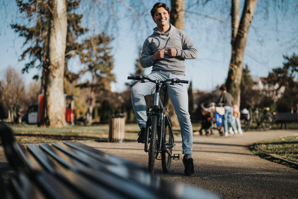 Ένας χαλαρός αρσενικός ποδηλάτης σταματά σε ένα παγκάκι σε ένα καλά φωτισμένο μονοπάτι που περιβάλλεται από δέντρα και οικογένειες σε ένα πάρκο της πόλης. - Φωτογραφία, εικόνα