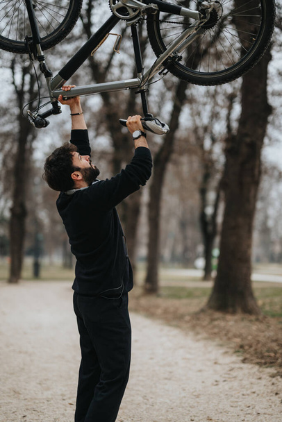 Бородатый бизнесмен в повседневной одежде поднимает велосипед над головой на парковой дорожке, демонстрируя силу и здоровый образ жизни. - Фото, изображение