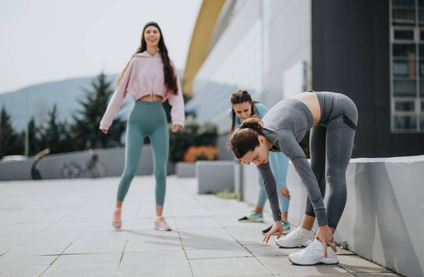 Trois jeunes femmes qui font des étirements sur un trottoir, se préparant pour un jogging dans la ville, avec un accent sur la forme physique et l'amitié. - Photo, image