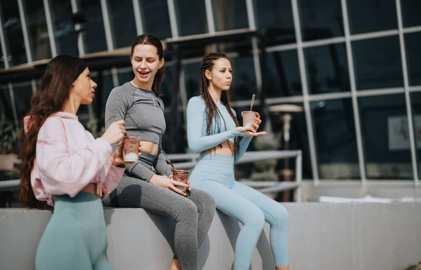 Trois femmes en tenue de sport assises à l'extérieur, sirotant des smoothies nutritionnels, et profitant de l'autre compagnie après une séance de fitness. - Photo, image