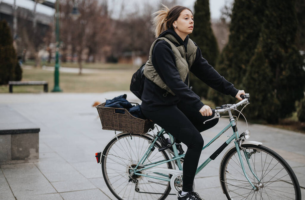 Jeune adulte concentré pédalant un vélo classique avec un panier en osier sur un sentier pavé dans un parc urbain, affichant un mode de vie actif et les transports urbains. - Photo, image
