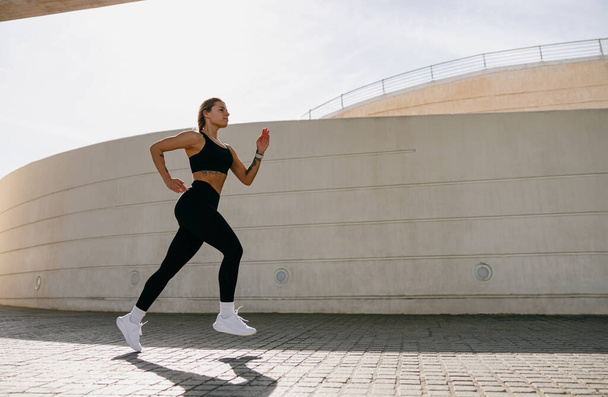 Fit αθλήτρια γυναίκα στο αθλητικό ντύσιμο τρέχει σε σύγχρονα κτίρια φόντο. Έννοια ενεργού τρόπου ζωής - Φωτογραφία, εικόνα