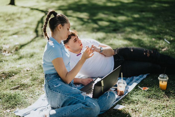 Двое молодых людей занимаются на свежем воздухе в солнечный день, пользуются ноутбуками и прохладительными напитками в парке. - Фото, изображение