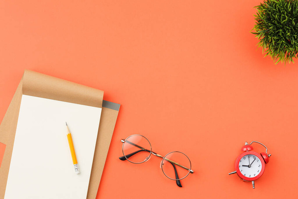 Craft notebook met witte lakens, rode wekker en potlood, glazen en groene bladeren van een plant in een pot op een oranje geïsoleerde achtergrond bovenaanzicht. - Foto, afbeelding