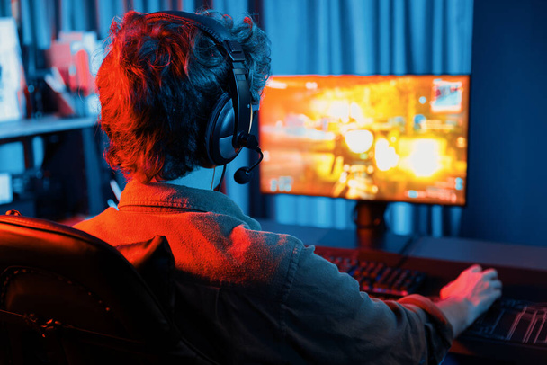 Kanał hosta młodego streamera gier, gracz zespołowy grający w gry wojenne strzelanie z multiplayerem na okręcie wojennym na ekranie komputera z obrazem z tyłu, noszenie słuchawki z mikrofonem w cyfrowym pokoju neonowym. Pędziwiatr. - Zdjęcie, obraz