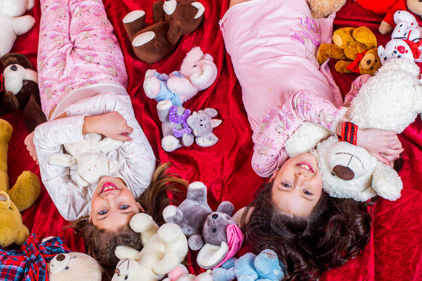 As irmãs de pijama deitam-se na cama, vista de cima. Crianças em pijama macio e quente brincando em casa. Meninas em pijama colorido segurar animais de pelúcia engraçados, infância, felicidade. - Foto, Imagem
