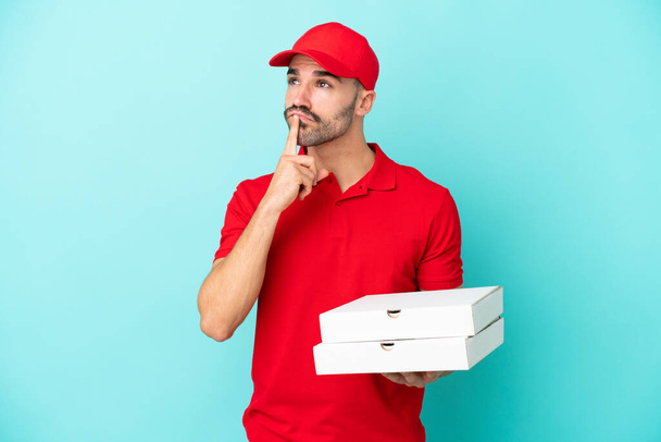 Dostawa mężczyzna kaukaski podnoszenia pudełka pizzy izolowane na niebieskim tle mając wątpliwości podczas patrząc w górę - Zdjęcie, obraz