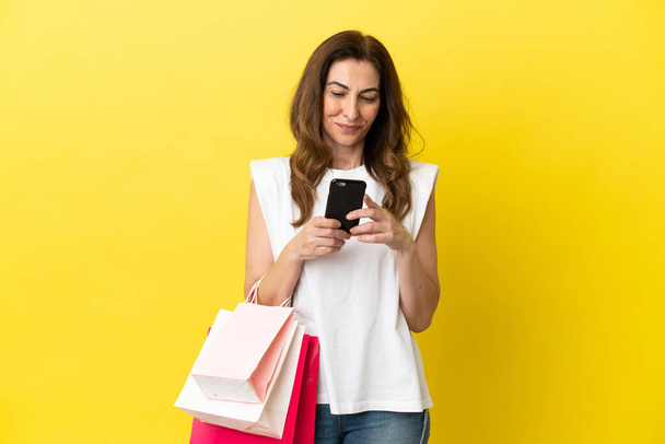 Mujer caucásica de mediana edad aislada sobre fondo amarillo sosteniendo bolsas de compras y escribiendo un mensaje con su teléfono celular a un amigo - Foto, imagen
