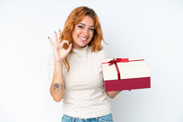 Young caucasian woman holding a gift iso0lated on white background showing ok sign with fingers - Photo, Image
