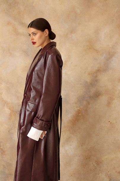 Portrait shooting of a stylish European girl in the studio. Dark hair and red lips. Model in a bodysuit and a long burgundy leather raincoat and sandals. Beige background. Fashion & Catalogs. Clothing trends for spring and autumn,trench - Photo, Image