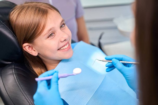 Hymyilevä tyttö istuu hammaslääkäreiden tuolissa, asiantuntija alkaa tutkia tyttöjen hampaita. - Valokuva, kuva