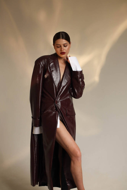 Portrait shooting of a stylish European girl in the studio. Dark hair and red lips. Model in a bodysuit and a long burgundy leather raincoat and sandals. Beige background. Fashion & Catalogs. Clothing trends for spring and autumn,trench - Photo, Image