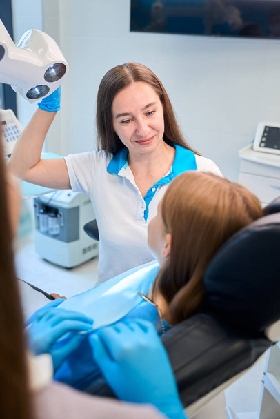 Dentista comienza a examinar a una paciente joven, la sudadera de las niñas está cubierta con una servilleta azul - Foto, imagen