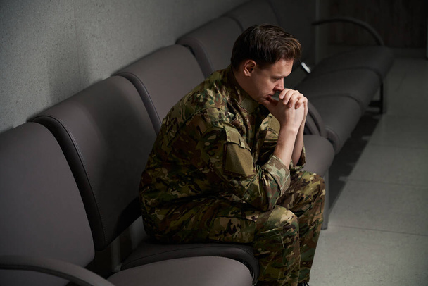 Dejected serviceman in camouflage uniform propping his chin with thumbs seated on leather sofa in waiting area - Photo, Image