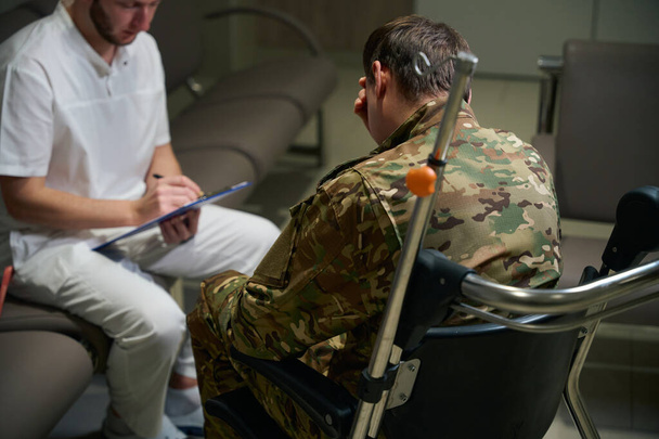 Ernsthafter Gesundheitsexperte macht sich Notizen mit Stift auf Klemmbrett in Anwesenheit eines Soldaten im Rollstuhl im Krankenhausflur - Foto, Bild