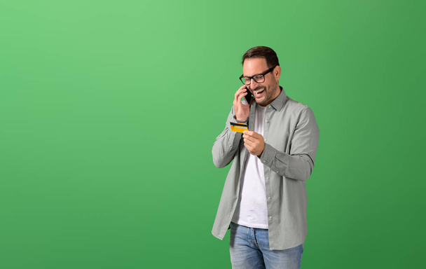 Χαρούμενος νεαρός άνδρας μοιράζονται τα στοιχεία της πιστωτικής κάρτας, ενώ μιλάμε για έξυπνο τηλέφωνο σε πράσινο φόντο - Φωτογραφία, εικόνα