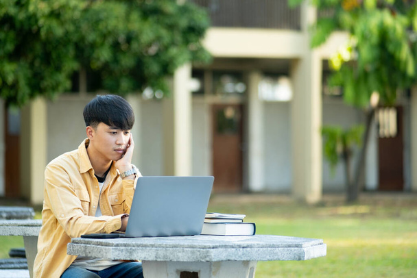 Un estudiante universitario se absorbe en el pensamiento mientras estudia en su computadora portátil, sentado en una mesa al aire libre rodeado de la tranquilidad del campus. - Foto, imagen