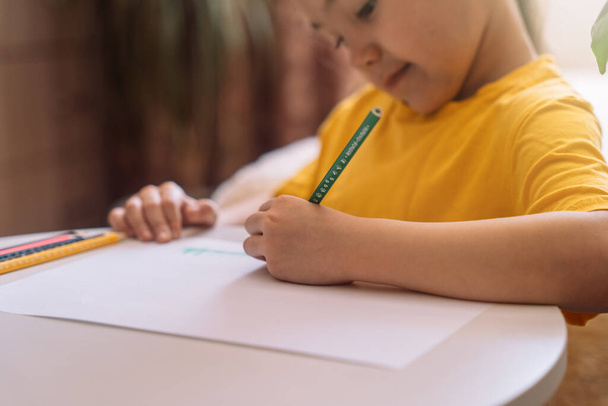 Un niño dibuja en la mesa con un lápiz verde sobre una hoja de papel. - Foto, Imagen