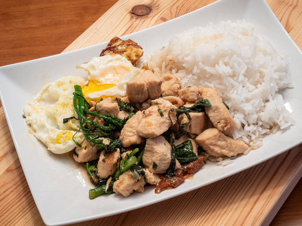 Indulge in the flavors of Thai basil chicken served with a perfectly fried egg, broccolini, and fragrant jasmine rice, elegantly presented on a white plate atop a rustic wooden board. - Photo, Image