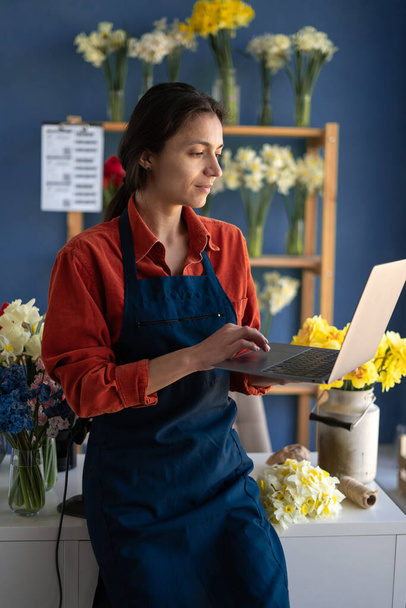 Portrait de femme fleuriste debout à sa boutique de fleurs et utilisant un ordinateur portable pour prendre des commandes pour son magasin. Espace de copie - Photo, image