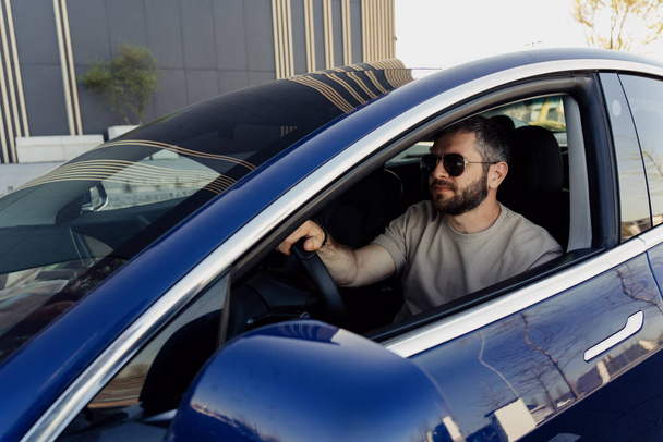 Ένας γενειοφόρος με γυαλιά ηλίου οδηγεί το μπλε ηλεκτρικό του αυτοκίνητο, αντανακλώντας ένα σύγχρονο, βιώσιμο τρόπο ζωής σε ένα αστικό περιβάλλον. - Φωτογραφία, εικόνα