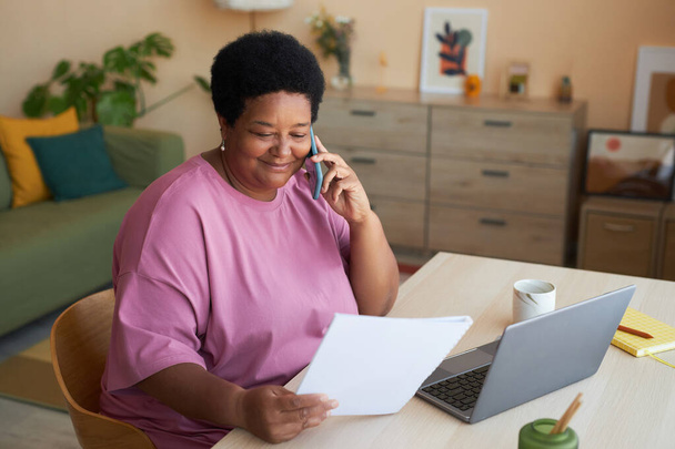Glimlachende oude zakenvrouw met financieel document praten met haar werkgever op mobiele telefoon terwijl zitten aan het bureau in de voorkant van de laptop - Foto, afbeelding