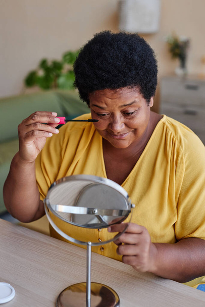 Afro-Amerikaanse oma in casualwear aanbrengen van make-up op haar gezicht en kijken in de spiegel terwijl zitten aan tafel in de voorkant van de camera thuis - Foto, afbeelding