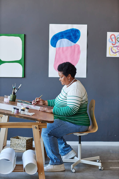 Lange shot van volwassen Afro-Amerikaanse vrouwelijke kunstenaar in casualwear creëren van nieuw schilderij terwijl zitten door houten bureau in de voorkant van de camera in de studio - Foto, afbeelding