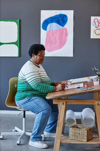 Серьезные творческие зрелая женщина в повседневной живописи наряд с гуашь на бумаге, сидя за столом в студии и создания новых произведений искусства - Фото, изображение