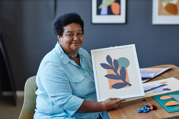 Creatief volwassen Afro-Amerikaanse vrouwelijke ontwerper met handgemaakte papieren applicatie in fotolijstje en kijken naar de camera in de studio - Foto, afbeelding