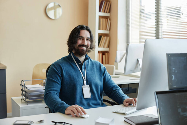 Joven gestor de TI sonriente en jersey azul usando la computadora y mirando a la cámara mientras está sentado en el lugar de trabajo y trabajando sobre un nuevo software - Foto, imagen