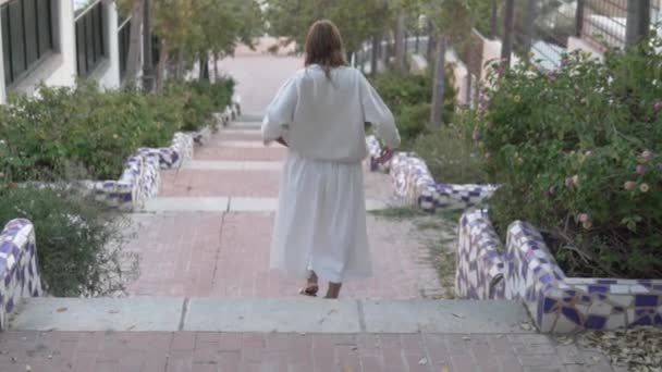 Eine Frau in einem weißen Kleid geht anmutig eine Treppe hinunter, die von sattgrünen Sträuchern und einer schimmernden Skulptur gesäumt ist, die vor dem Hintergrund eines baumbestandenen Bürgersteigs und Wasserspiels steht. - Filmmaterial, Video