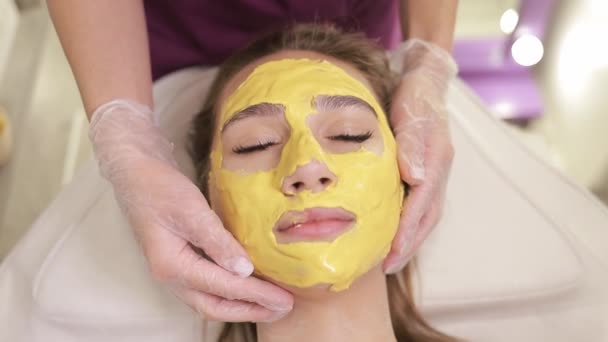 La mujer se está poniendo una mascarilla amarilla en su piel en el spa, cubriendo su cara de frente a barbilla. Ella tiene una sonrisa en su cara mientras la máscara se aplica suavemente con una mano - Metraje, vídeo