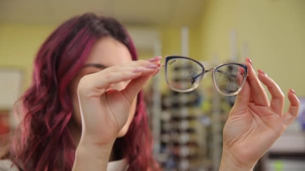 Ein Mädchen mit schönen roten lockigen Haaren wählt eine Brille, um ihre Sehkraft zu verbessern. Sehkorrektur. Auswahl professioneller Brillen im Optikgeschäft.  - Filmmaterial, Video