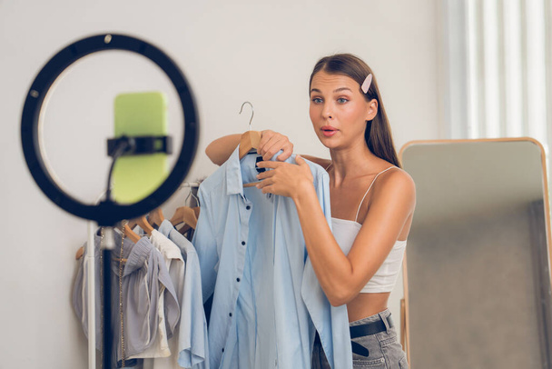 Young woman making fashion video content for social media. Blogger smiles to camera using selfie stick and light ring while making persuasive online clothing sell to audience or followers. Unveiling - Photo, Image