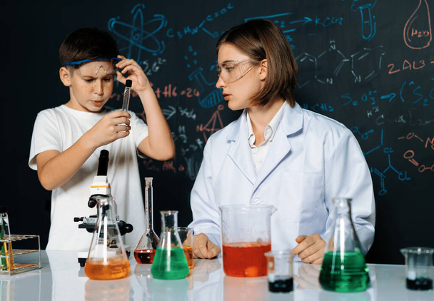 Nauczyciel wspiera ucznia w laboratorium. Uczeń i nauczyciel stoją i eksperymentują na chemii w klasie STEM używając cieczy w szklanym pojemniku. Roztwór mieszający instruktora. Erudycja. - Zdjęcie, obraz
