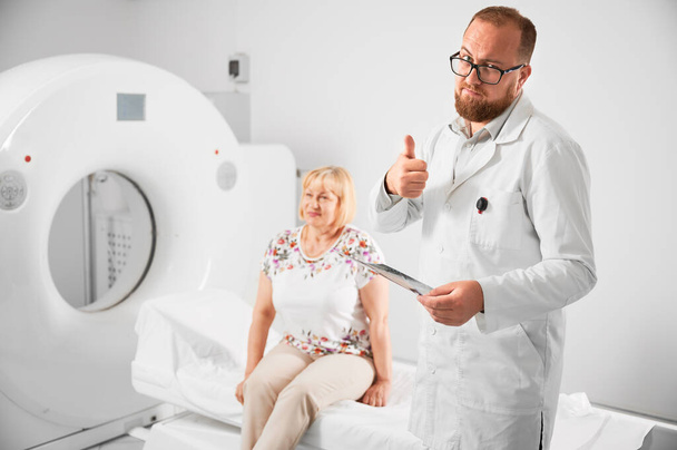 Medische computertomografie of MRI scanner. Dokter met MRI-resultaten, met duimen omhoog gebaar. Een vrouw die op de bank zit. Begrip geneeskunde, gezondheidszorg en moderne diagnostiek. - Foto, afbeelding