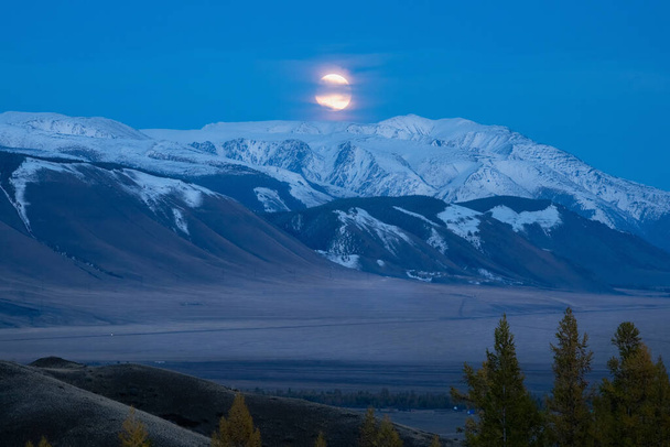 Csodálatos misztikus holdfényes éjszaka egy hegyi völgyben. Nagy hold egy havas hegy felett. Arany telihold emelkedik a távoli hegyek felett a kék órában. Tökéletes kép a falra, képernyőre. Forgatókönyv - Fotó, kép