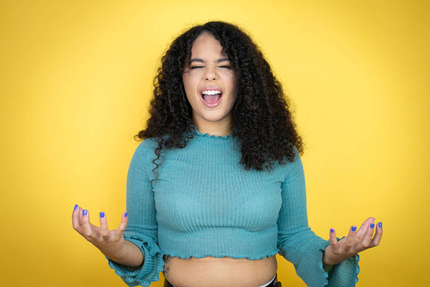 Afro-Amerikaanse vrouw draagt casual trui over gele achtergrond gek en gek schreeuwen en schreeuwen met agressieve uitdrukking en armen omhoog. Frustratieconcept. - Foto, afbeelding
