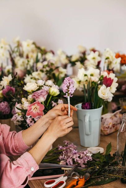 Egy rózsaszín inges személy orgona jácintokat rendez, körülvéve különféle friss virágokkal és virágdíszítő szerszámokkal. - Fotó, kép