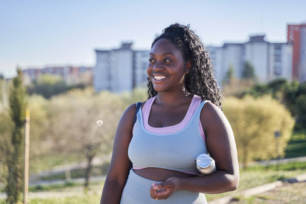 молода афроамериканка, що проходить через парк у спортивному одязі, тримає пляшку води - Фото, зображення