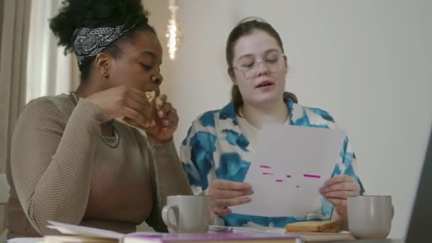 Pas w górę ujęcia dwóch wielorasowych dziewcząt po dwudziestce odrabiających razem pracę domową przy stole w nowoczesnym mieszkaniu - Materiał filmowy, wideo