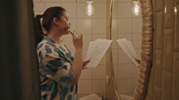 Seitenansicht mittlere Aufnahme einer jungen kaukasischen Plus-Size-Frau, die vor einem Badezimmerspiegel mit Studienmaterialien auf Papierbögen in den Händen steht, Gua-sha-Gesichtsmassage macht und sich auf die Prüfung vorbereitet - Filmmaterial, Video